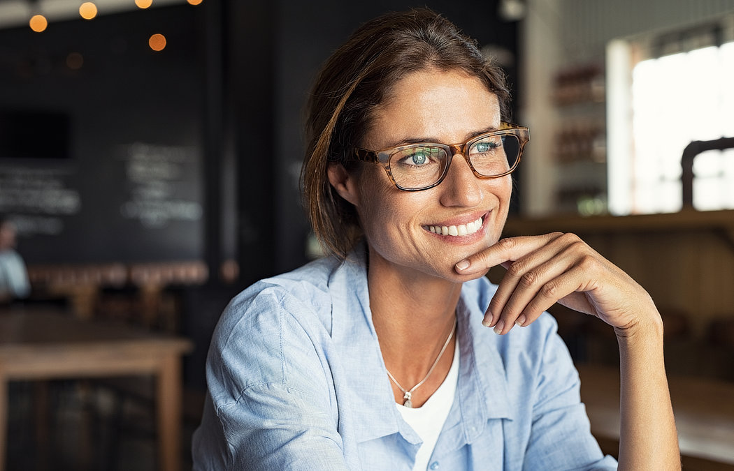 A woman wearing glasses and a light blue shirt smiles while resting her chin on her hand in a cafe setting., Lawrenceville, GA