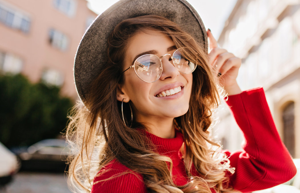 Woman in a red sweater smiles while touching her hat. She is wearing round glasses and hoop earrings, standing outdoors with buildings in the background., Lawrenceville, GA